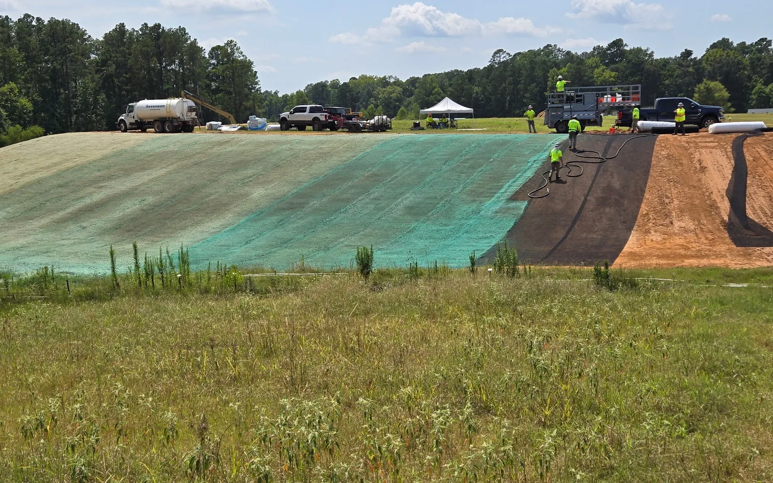 Remediation slope mid-installation, striped with green hydromulch, dark turf reinforcement mats, and bare earth, with crew and trucks on the crest